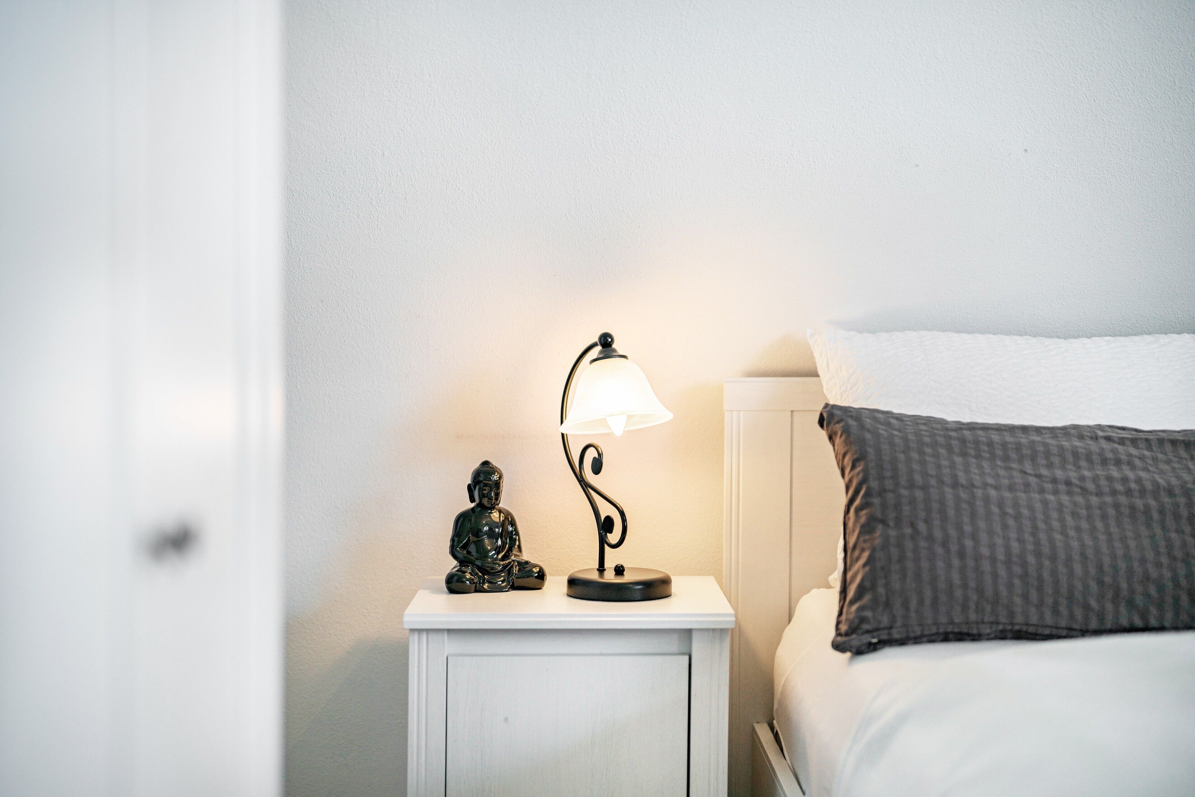 Een witte nachtkastje met een lamp en een Boeddhabeeld, een bed met een grijs kussen en een witte muur.