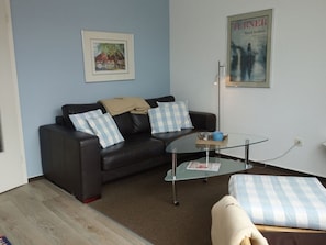Un salon avec un canapé en cuir foncé, une table basse en verre et un pouf à carreaux bleus et blancs. Un tableau encadré est accroché au mur, et une affiche est posée sur le mur opposé.