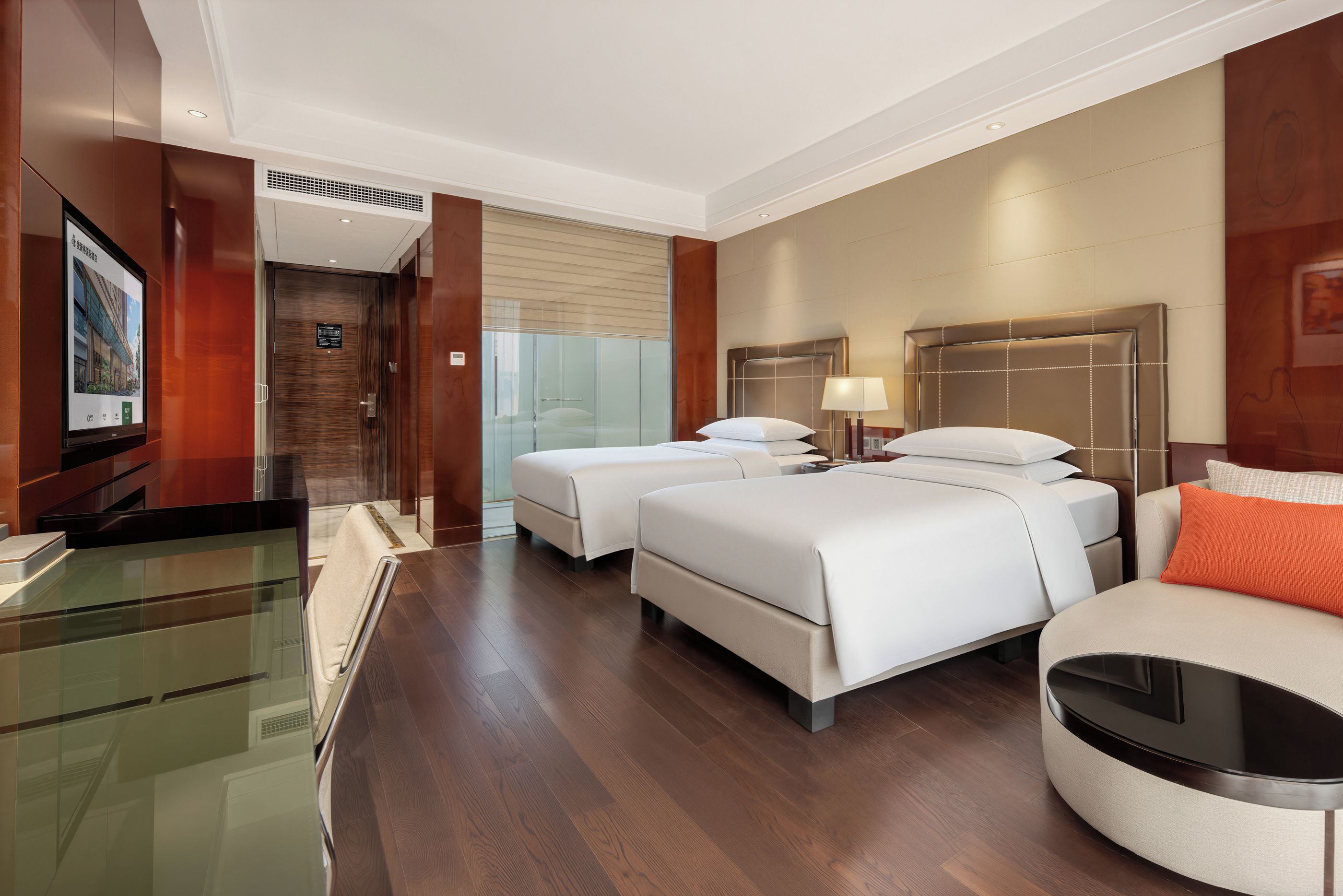 Quarto de hotel com escrivaninha de vidro, duas camas, um sofá e uma televisão na parede.
