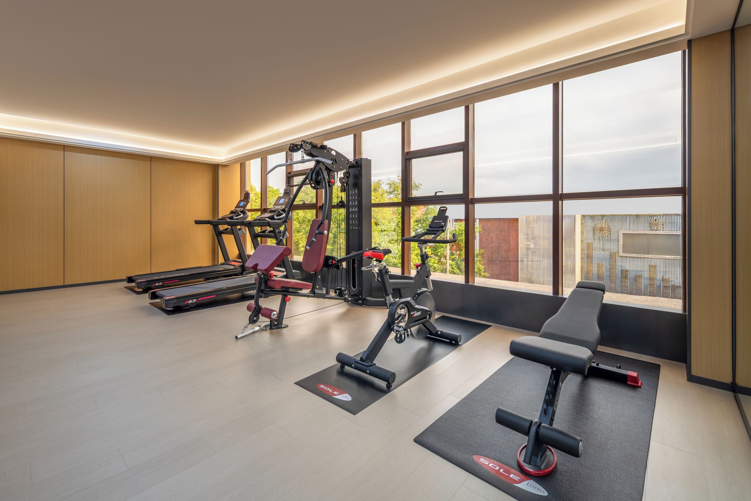 A modern gym with treadmills, weight machines, and a bench. Large windows provide natural light and views of the outdoors.