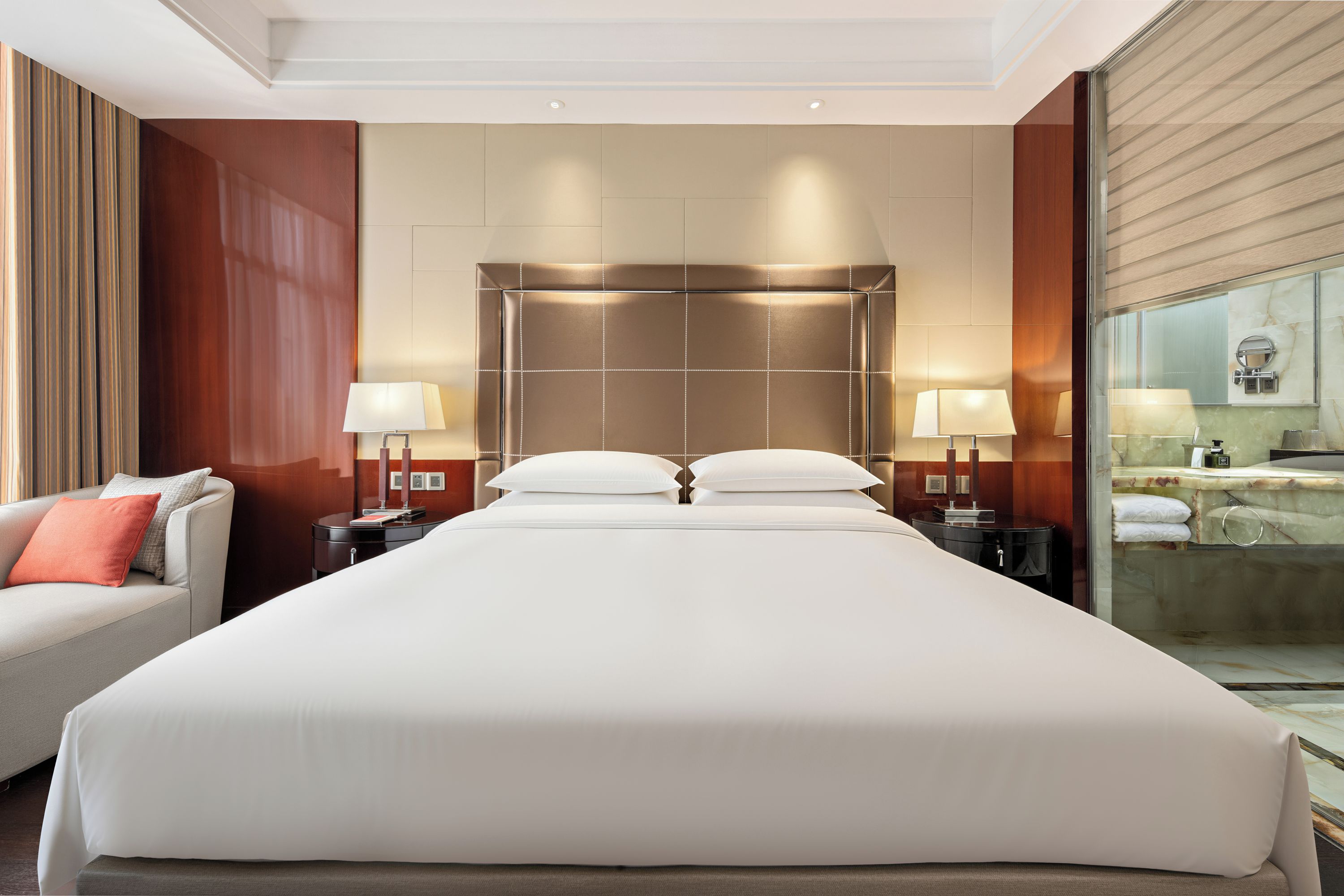 A hotel room with a large bed, two bedside tables, a sofa, and a bathroom with a glass partition.