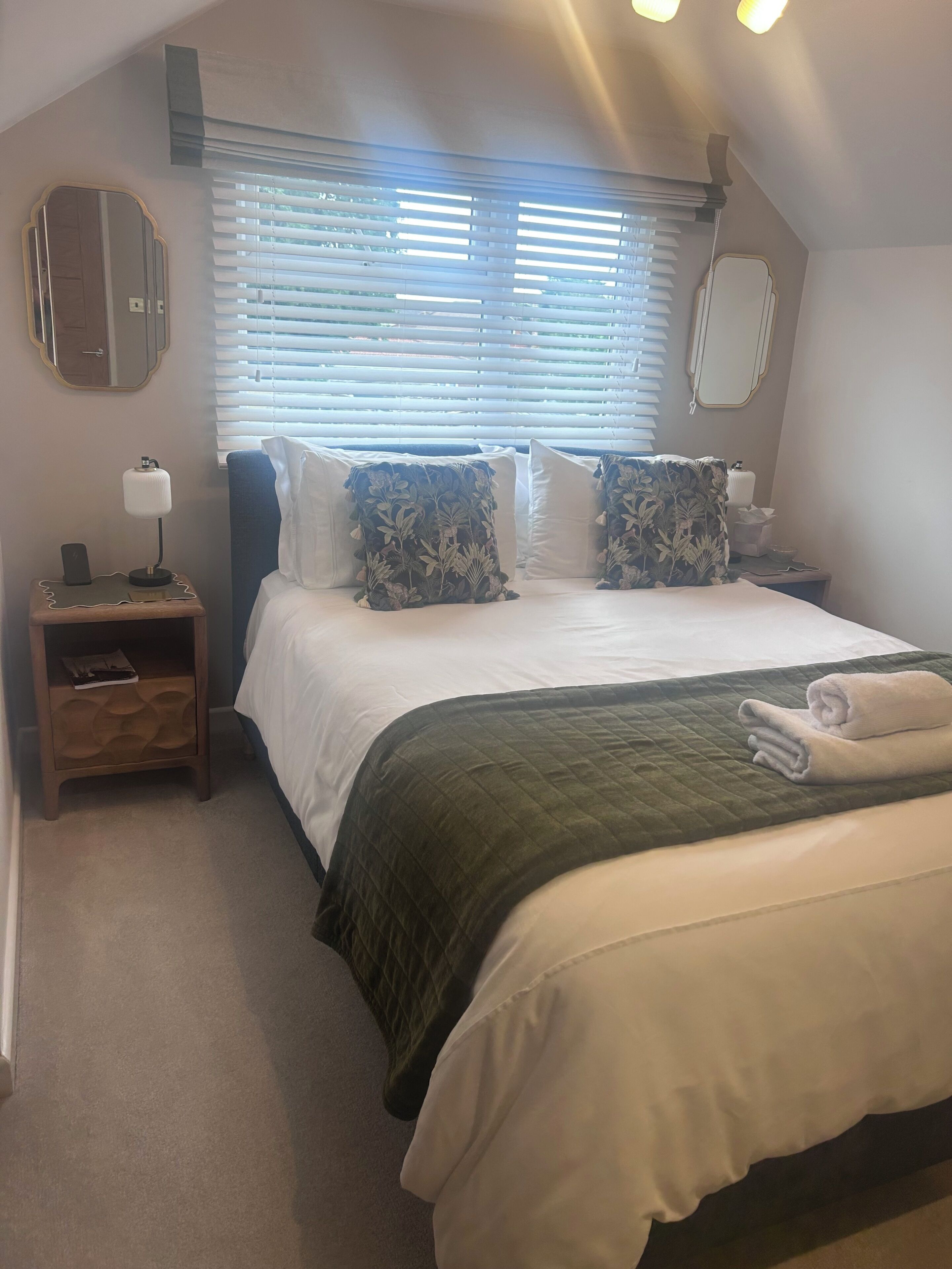 Ein Schlafzimmer mit einem Bett, einem Nachttisch, einer Lampe, einem Spiegel und einem Fenster mit Jalousien.