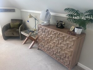 Sala de espera con una silla, una mesa con cubierta de vidrio y controles remotos, una lámpara, un ventilador, un teléfono antiguo y una planta en maceta.