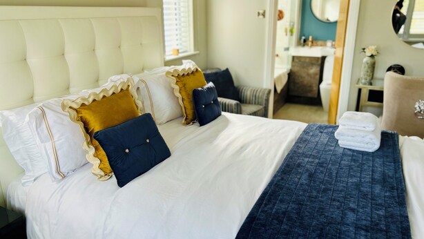 Una cama bien hecha con almohadas blancas y azul oscuro, una manta azul y una mesita con toallas.