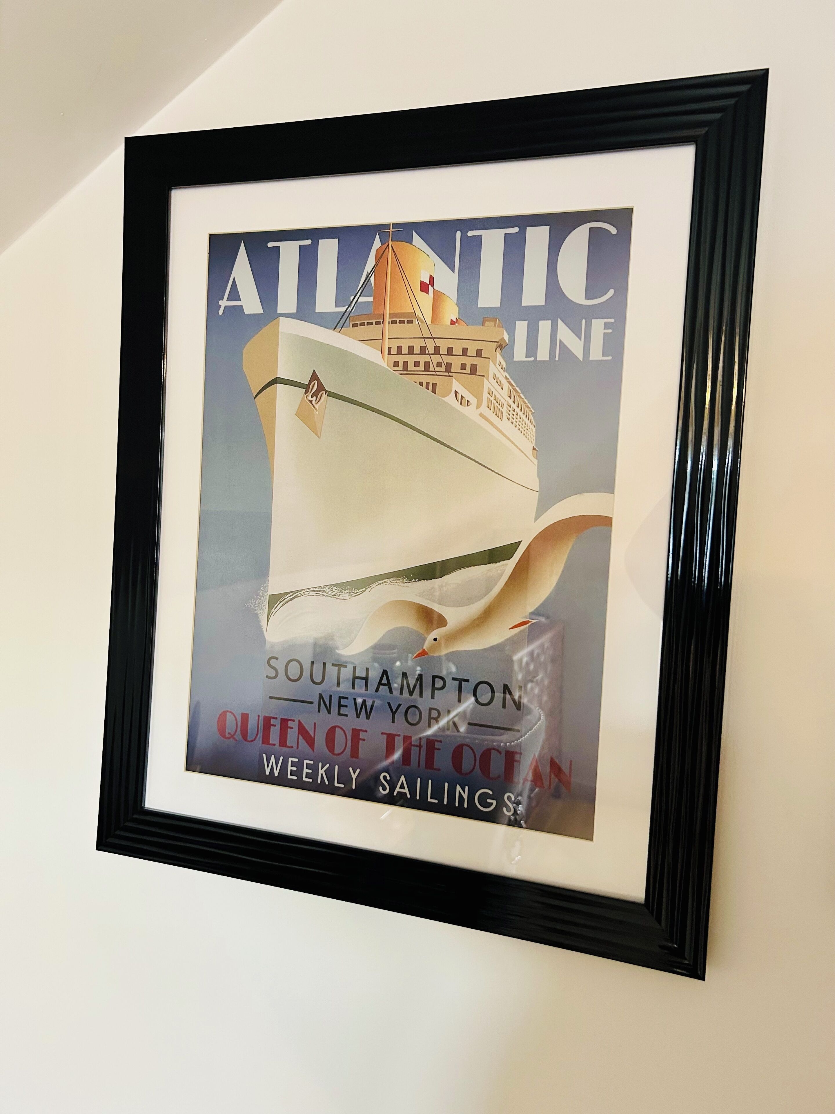 Une affiche encadrée représentant un navire de la compagnie Atlantic Line, avec le texte suivant : « Southampton – New York – Queen of the Ocean – Traversées hebdomadaires ».