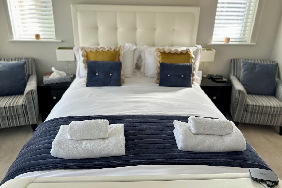 Habitación de hotel con una cama grande, dos mesitas de noche, una silla y una ventana con persianas.