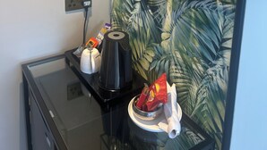 Un área de cocina con cafetera, hervidor y microondas en la encimera. Hay una estantería con tazas y un objeto decorativo.