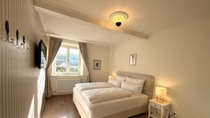 Une chambre à coucher avec un grand lit, une table de chevet avec une lampe, une fenêtre avec des rideaux, un radiateur et un téléviseur fixé au mur.