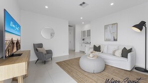 Una sala moderna con televisor, un sillón gris, un sofá blanco, una mesa de centro redonda y una cocina al fondo.
