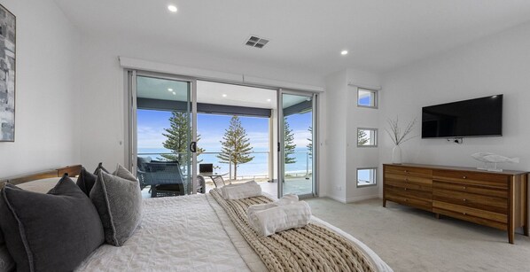 Une chambre moderne avec un grand lit, une commode en bois, un téléviseur à écran plat et un balcon offrant une vue sur l’océan et les arbres.