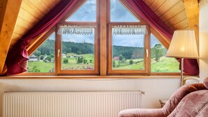 Une pièce avec un plafond en pente, deux fenêtres avec des rideaux, un radiateur et un fauteuil à motifs.