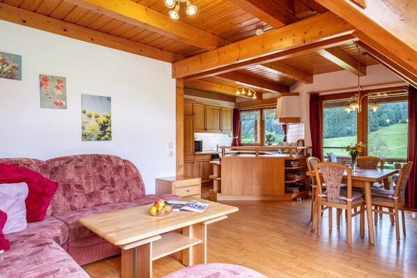 A cozy living room with a sofa, a wooden dining table with chairs, a kitchen area, and a view of the outdoors.