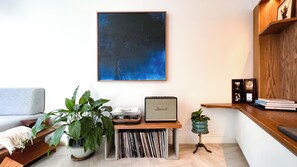 A living room with a large abstract painting, a Marshall amplifier, a plant, and a wooden shelf with books and photos.