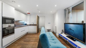 Una cocina moderna con armarios blancos, horno empotrado y microondas. En la sala de estar, un sofá azul, un televisor de pantalla plana y una ventana con cortinas.