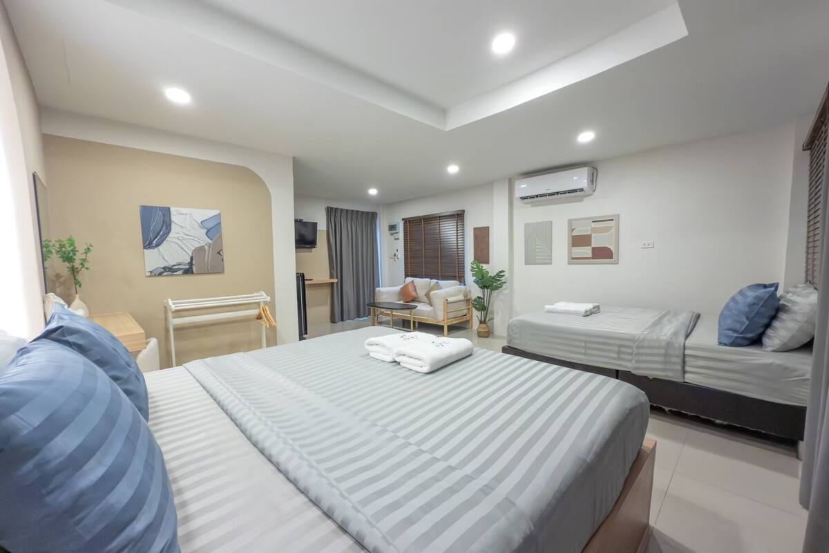 A modern hotel room with a large bed, a smaller bed, a sofa, a TV, and an air conditioning unit.