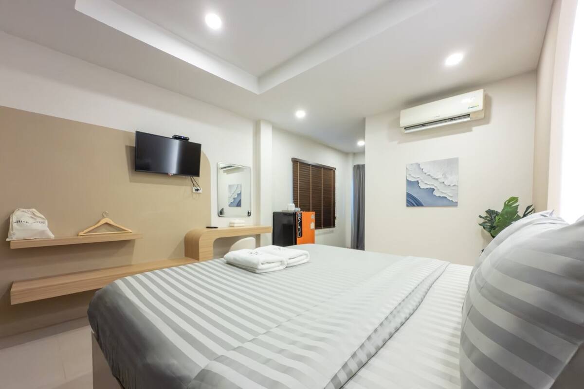 A modern hotel room with a bed, a desk, a chair, a TV, an air conditioner, and a plant.