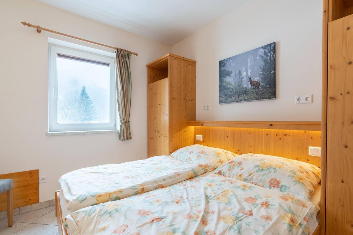 A bedroom with two beds, a wooden wardrobe, a window with curtains, a painting of a deer, and a bench.