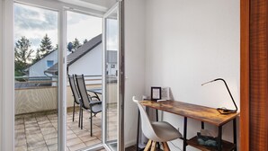 Een kleine kamer met een schuifdeur van glas naar een balkon, een houten bureau met een stoel en een klein plankje.