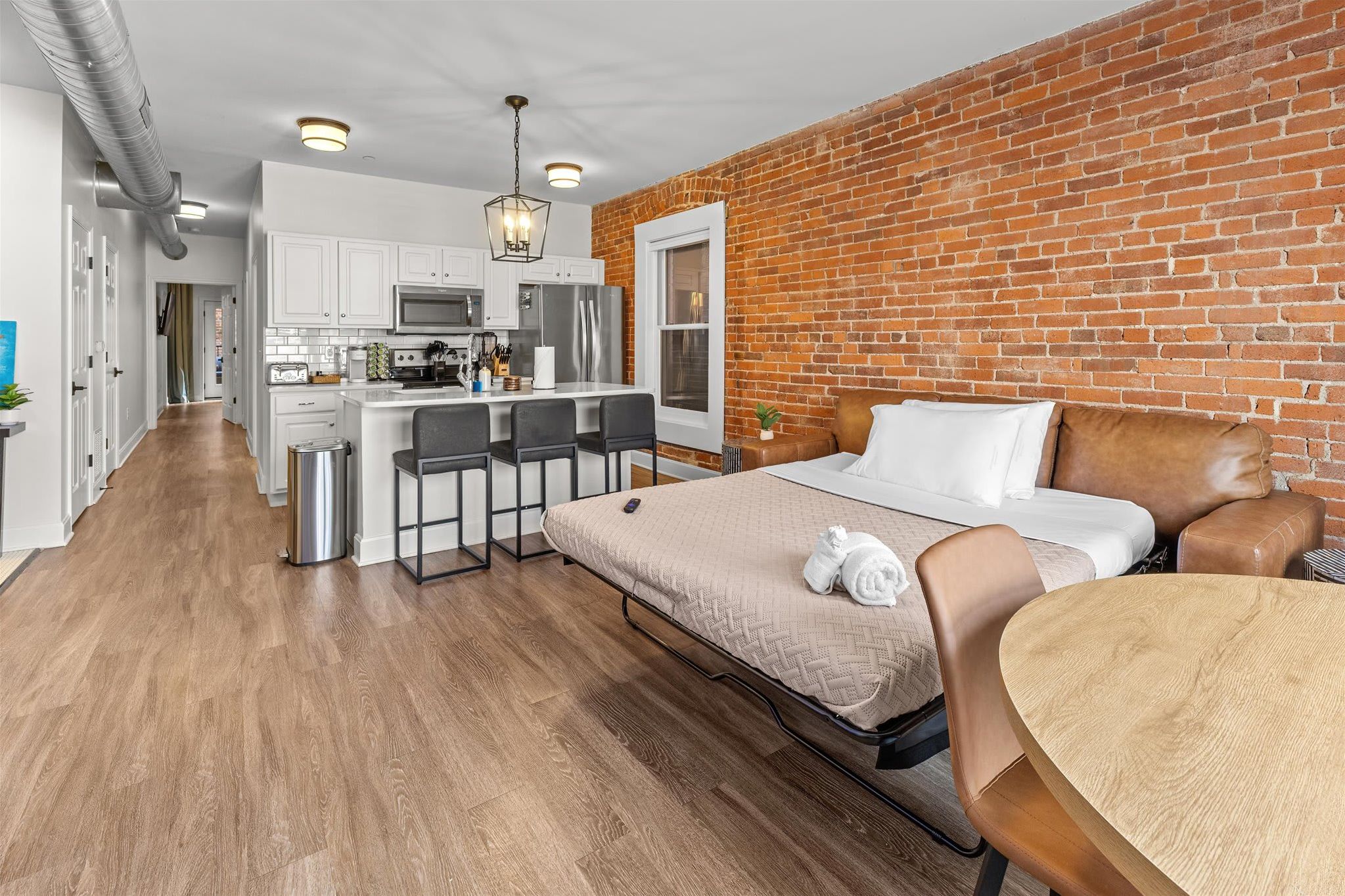 A compact hotel room with a bed, a small dining table, a kitchen area, and a brick wall.