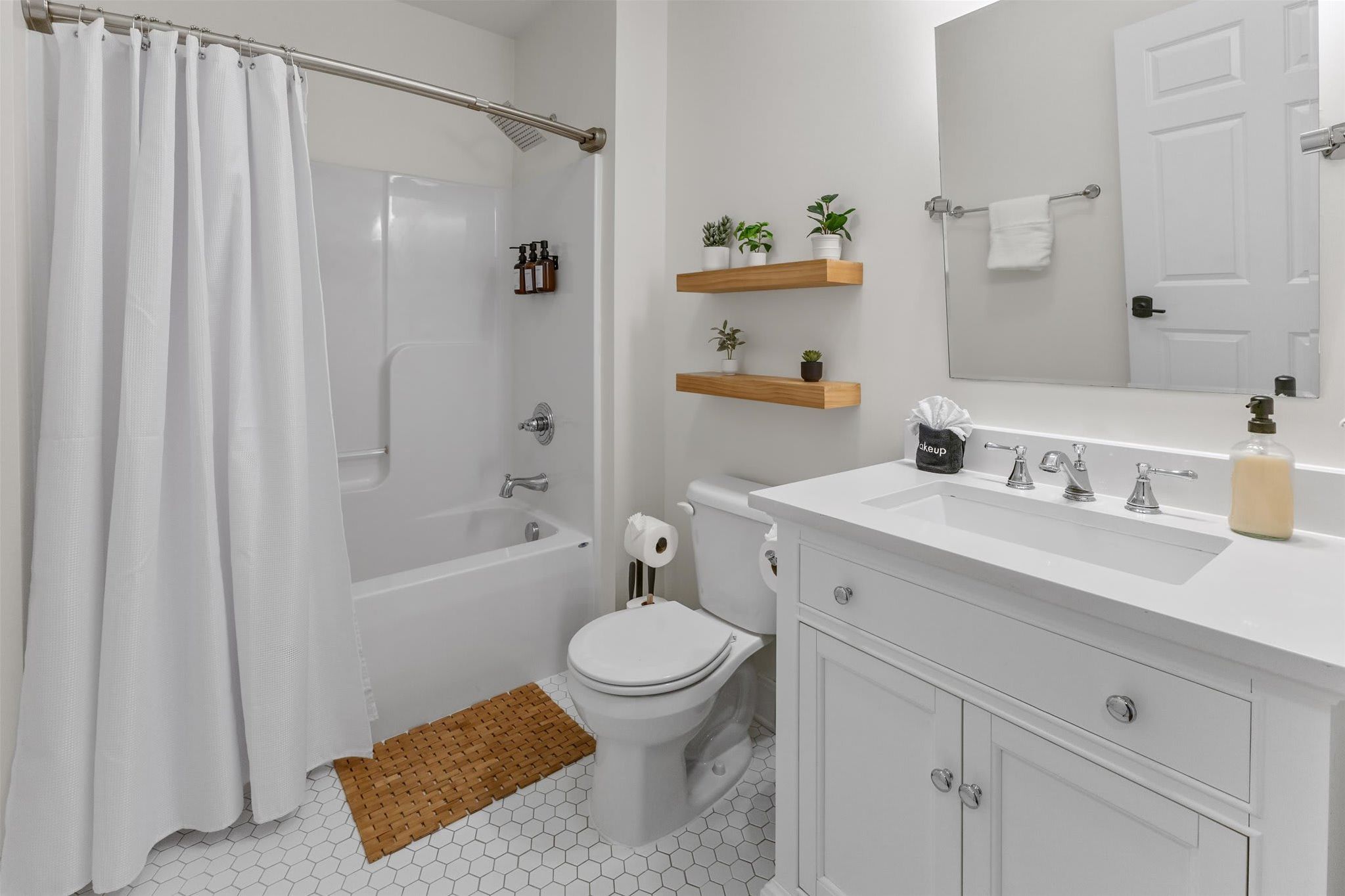 A bathroom with a white bathtub, toilet, and sink. There are wooden shelves with plants, a white shower curtain, and a hexagonal floor pattern.