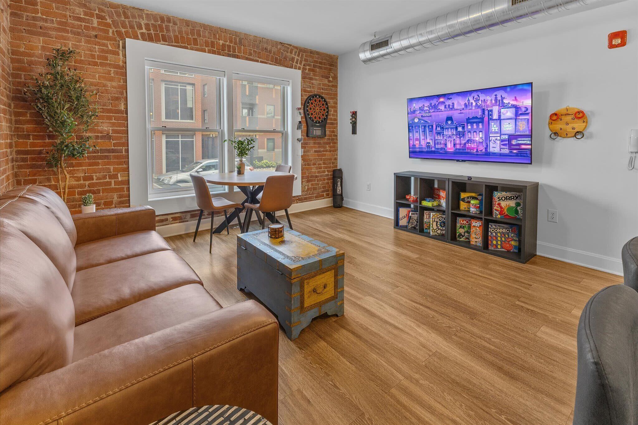 Een moderne woonkamer met een bakstenen muur, een houten vloer, een bruine leren bank, een salontafel, een televisie en een boekenkast met gezelschapsspellen.