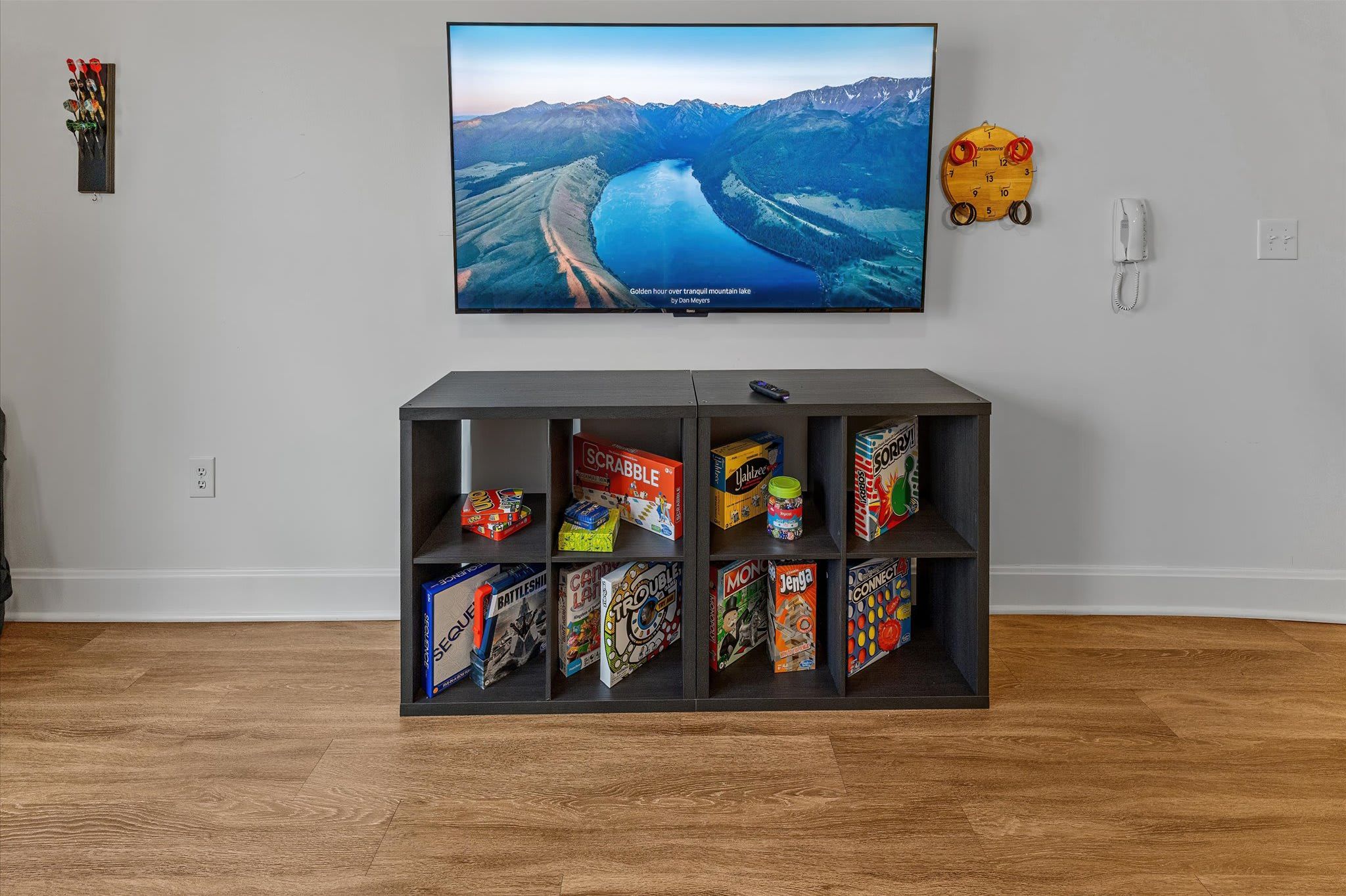 A living room with a flat-screen TV mounted on the wall, displaying a scenic landscape. Below the TV, there is a shelf with various board games.
