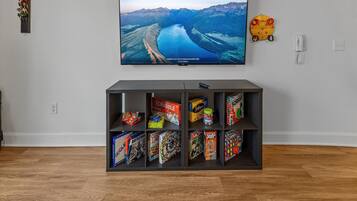 A living room with a flat-screen TV mounted on the wall, displaying a scenic landscape. Below the TV, there is a shelf with various board games.