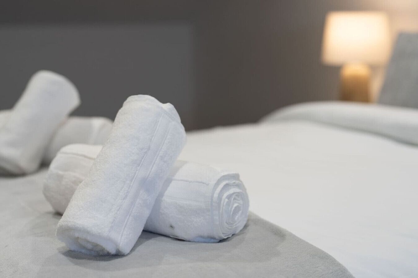 A neatly made bed with white linens and a rolled towel on a gray headboard.