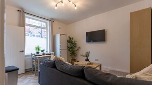 Un espacio habitable compacto con sofá, mesa de comedor, pequeña cocina y televisor fijado a la pared.