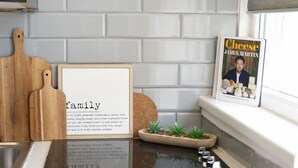 Una encimera de cocina con un marco que contiene una cita familiar, una tabla de cortar de madera, una pequeña planta en maceta, una revista titulada 'Cheese' de James Martin, y una ventana con vistas al exterior.