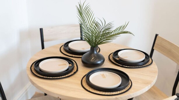 Ein runder Holztisch mit schwarzen Untersetzern und weißen Tellern, umgeben von Holzstühlen.