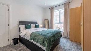 Ein Schlafzimmer mit einem Bett, einem Nachttisch, einer Lampe, einem Fenster mit Vorhängen und einer Tür.