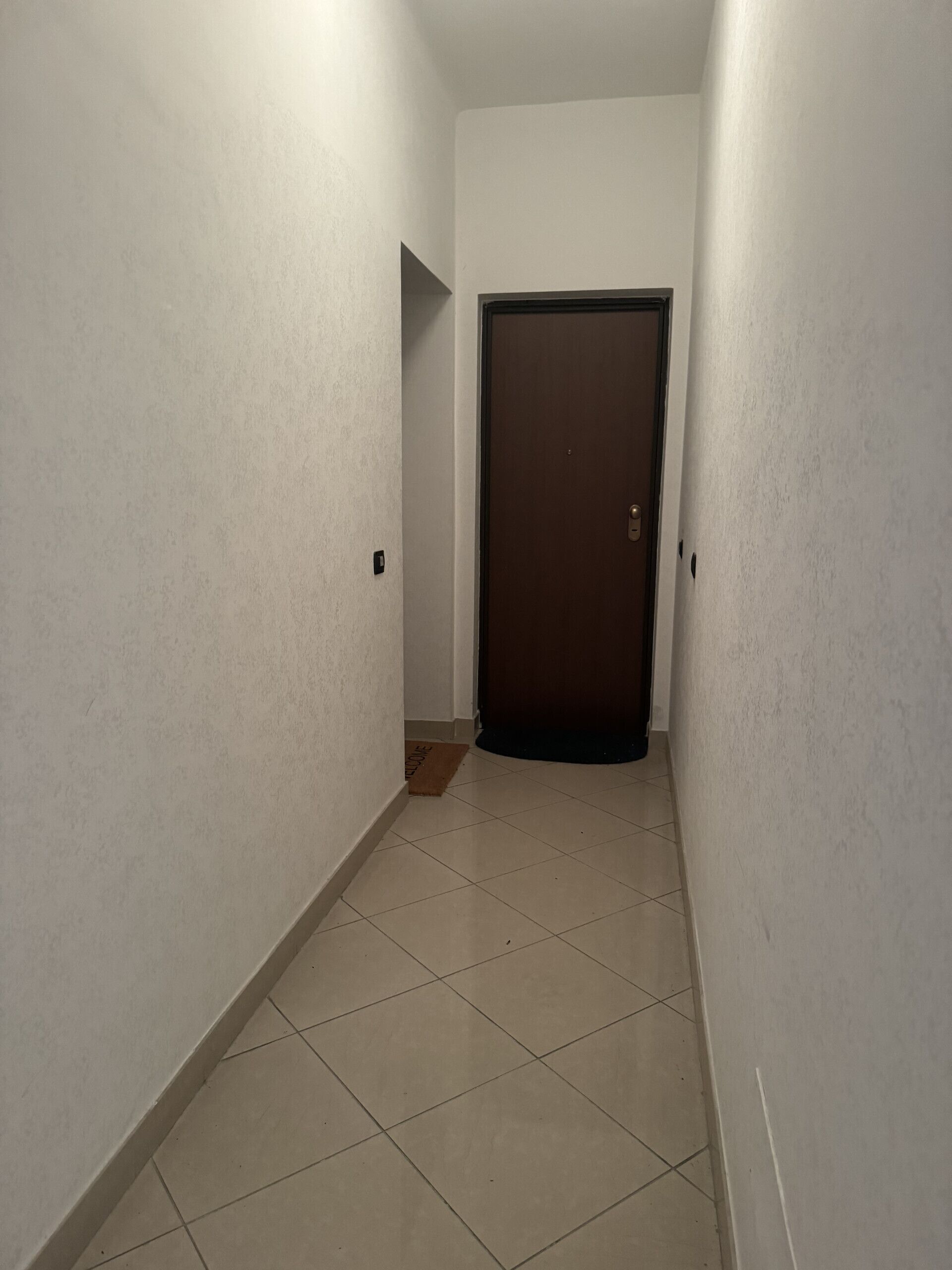 Un pasillo estrecho con paredes blancas y suelo de baldosas. Se ve una puerta al final del pasillo.