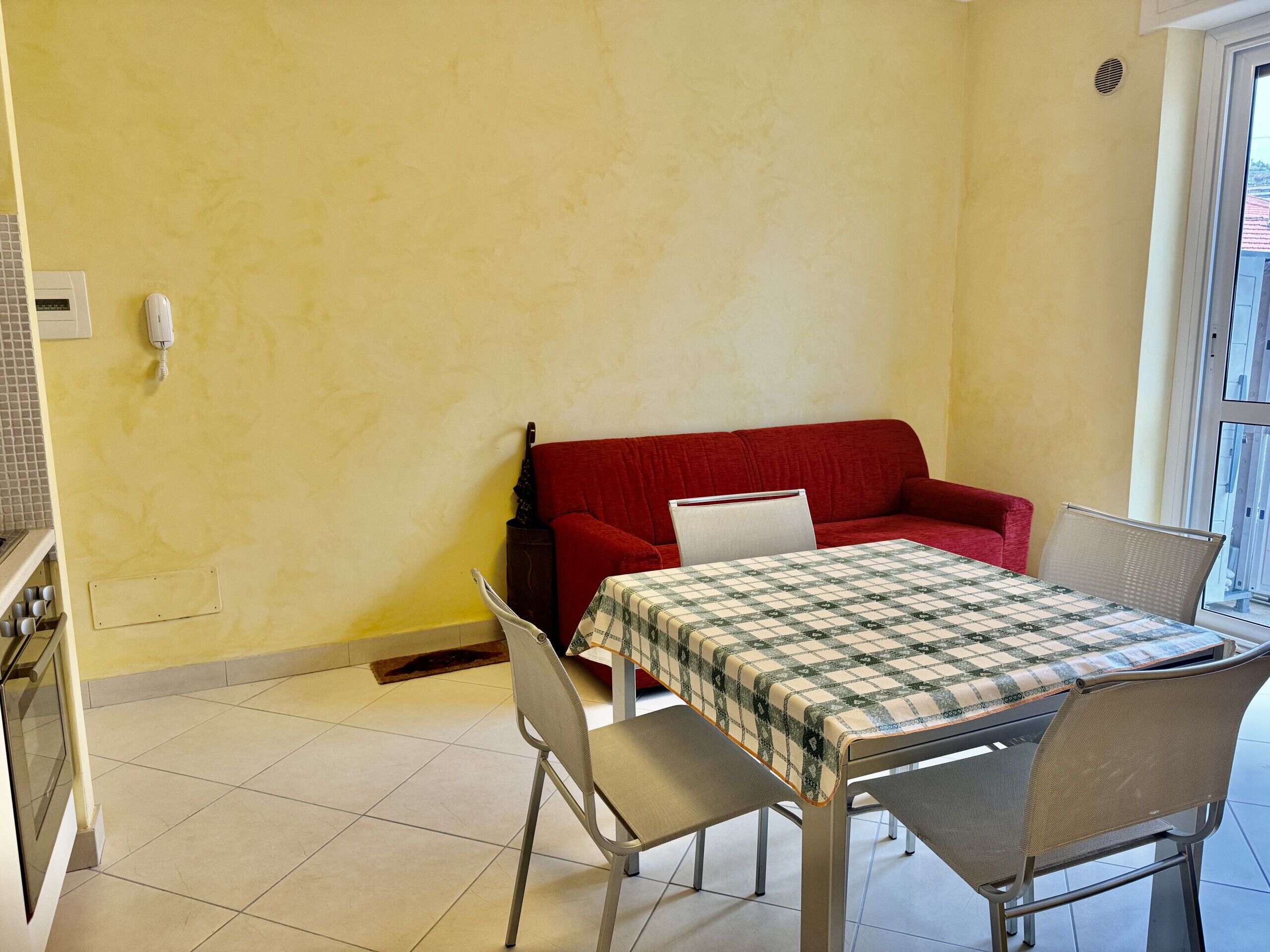 A compact living space with a red sofa, a dining table with a checkered tablecloth, and a kitchen area with a stove.