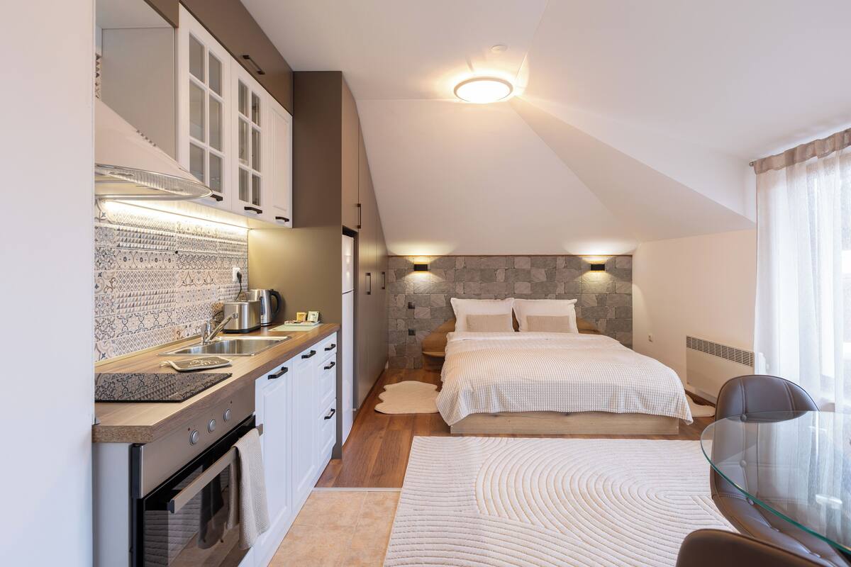 A compact bedroom with a stone accent wall, a bed with white bedding, a glass table, and a kitchenette with a stove and sink.