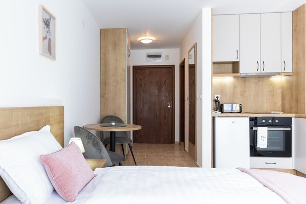 Quarto de hotel compacto com cama, mesa de cabeceira, cadeira, pequena mesa e cozinha compacta.