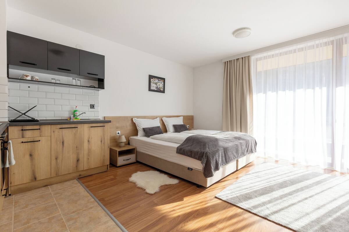 A modern bedroom with a wooden bed, a small kitchen area, and a large window with curtains.