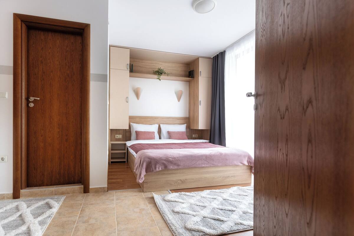 A bedroom with a wooden door, a bed with a headboard, a bedside table, and a nightstand.