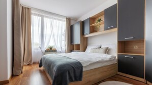 A modern bedroom with a wooden bed, built-in dark cabinets, and a window with sheer curtains.