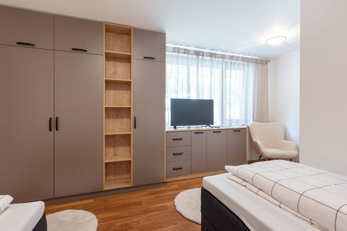 A bedroom with a bed, a TV, a chair, a wardrobe, and a desk.