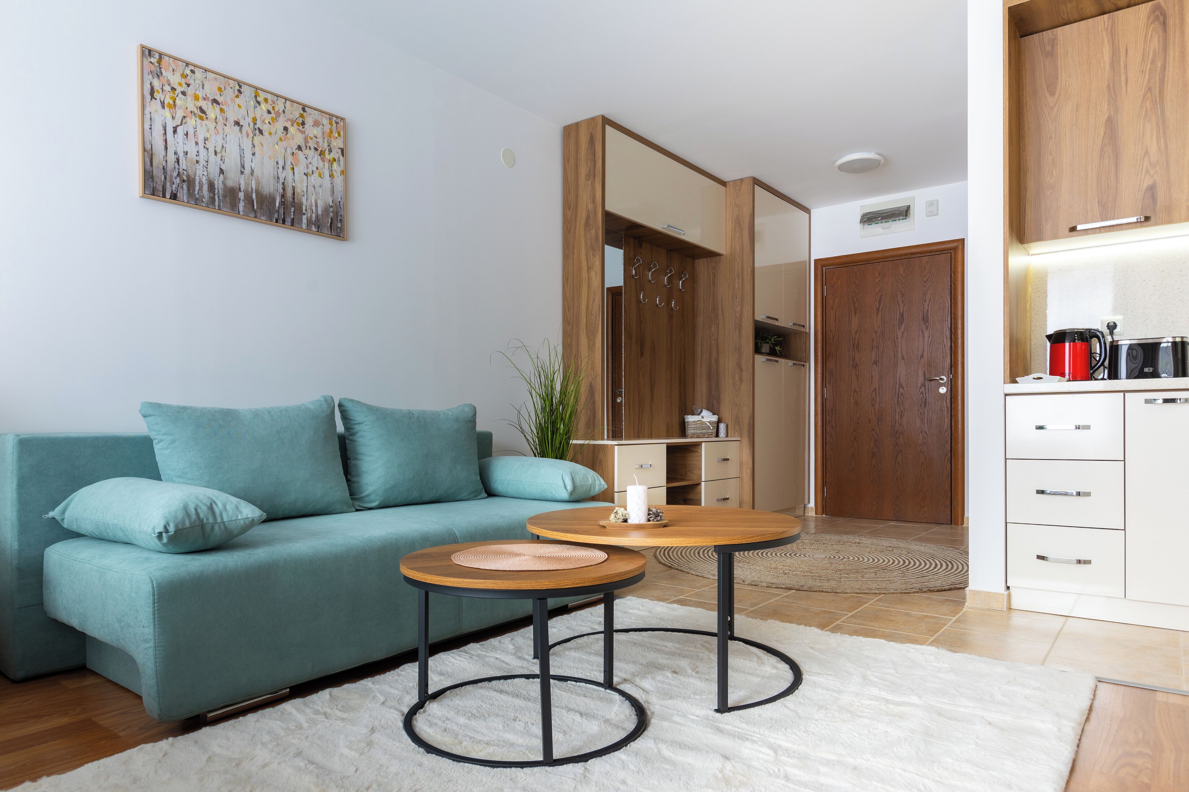 A modern living room with a teal sofa, two round tables, a white rug, and a kitchen area with wooden cabinets and a red kettle.