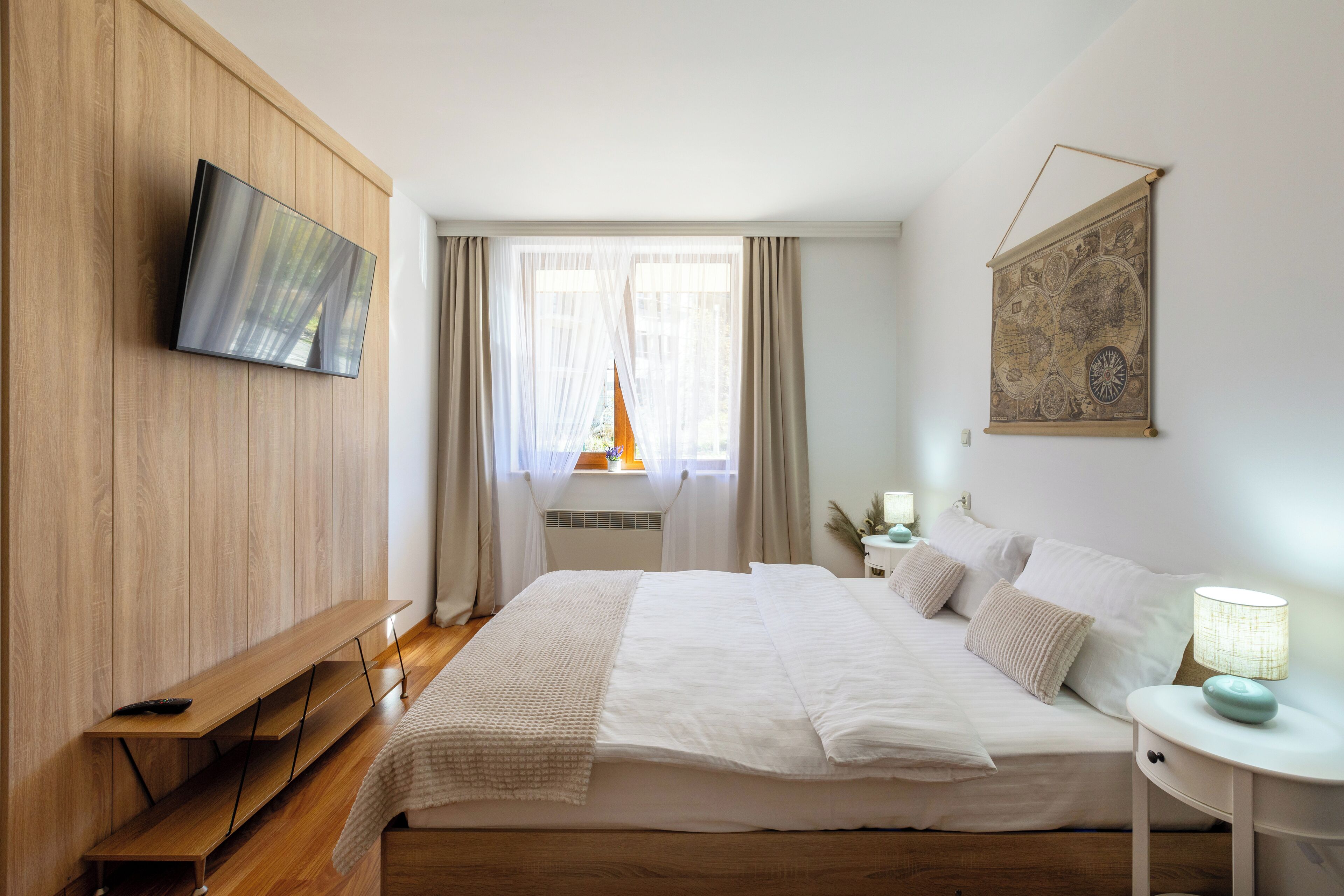 A bedroom with a bed, a TV mounted on the wall, a map on the wall, and a window with curtains.