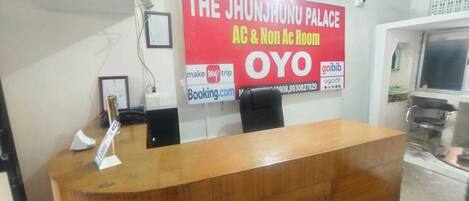 A reception desk with a wooden finish, a framed certificate on the wall, and a large promotional banner for The Jhunjhunu Palace AC & Non AC Rooms.
