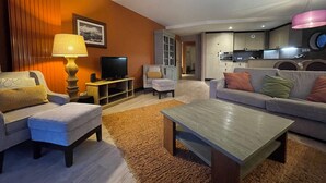 A modern living room with a television, sofa, and coffee table. A kitchen is visible in the background.