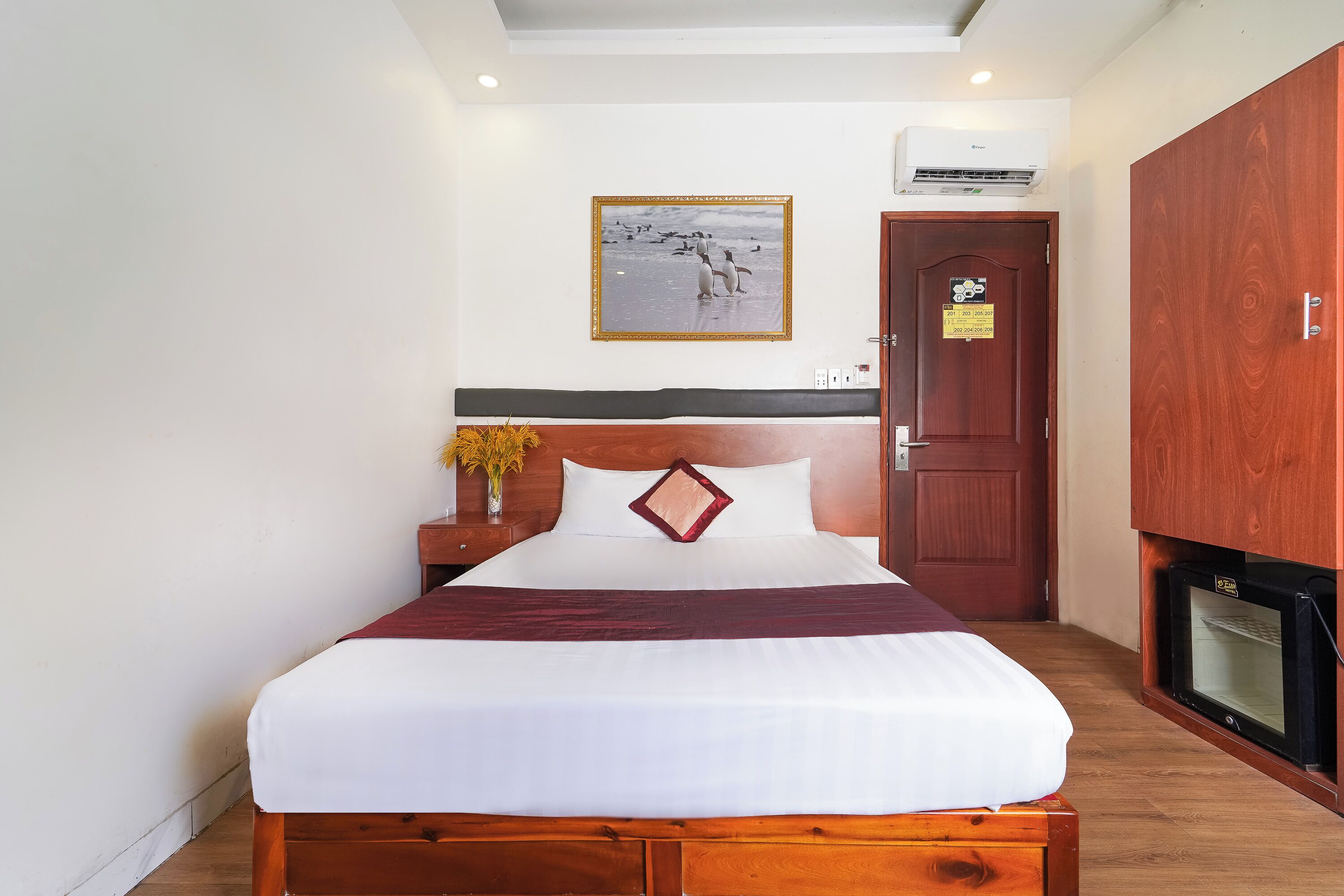 Un lit bien fait, avec du linge de lit blanc et un coussin rouge, une tête de lit en bois, un tableau encadré au mur, une armoire en bois et un petit téléviseur noir posé sur le sol.