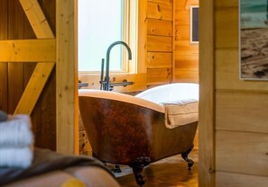 A wooden cabin interior with a clawfoot bathtub, a window, and a wooden door.