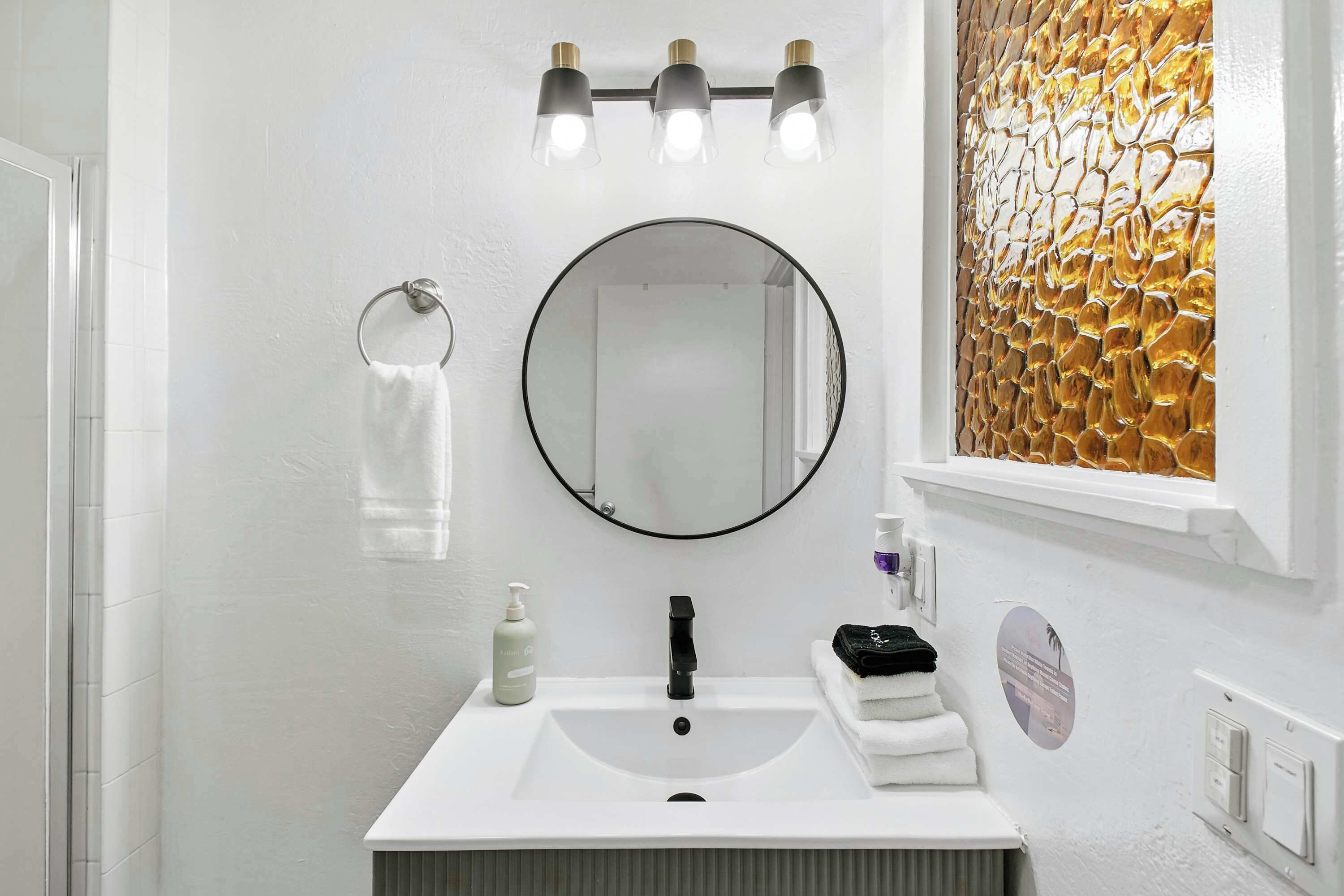 Un baño con un lavamanos blanco, un espejo redondo y un perchero. Hay tres luces sobre el espejo y un panel decorativo a la derecha.