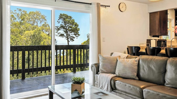 A living room with a sliding glass door to a balcony, a sofa, a coffee table, and a kitchen area with a clock on the wall. - @ Marbella Lane - Natural Bayview Escape Near SFO (Daly City)