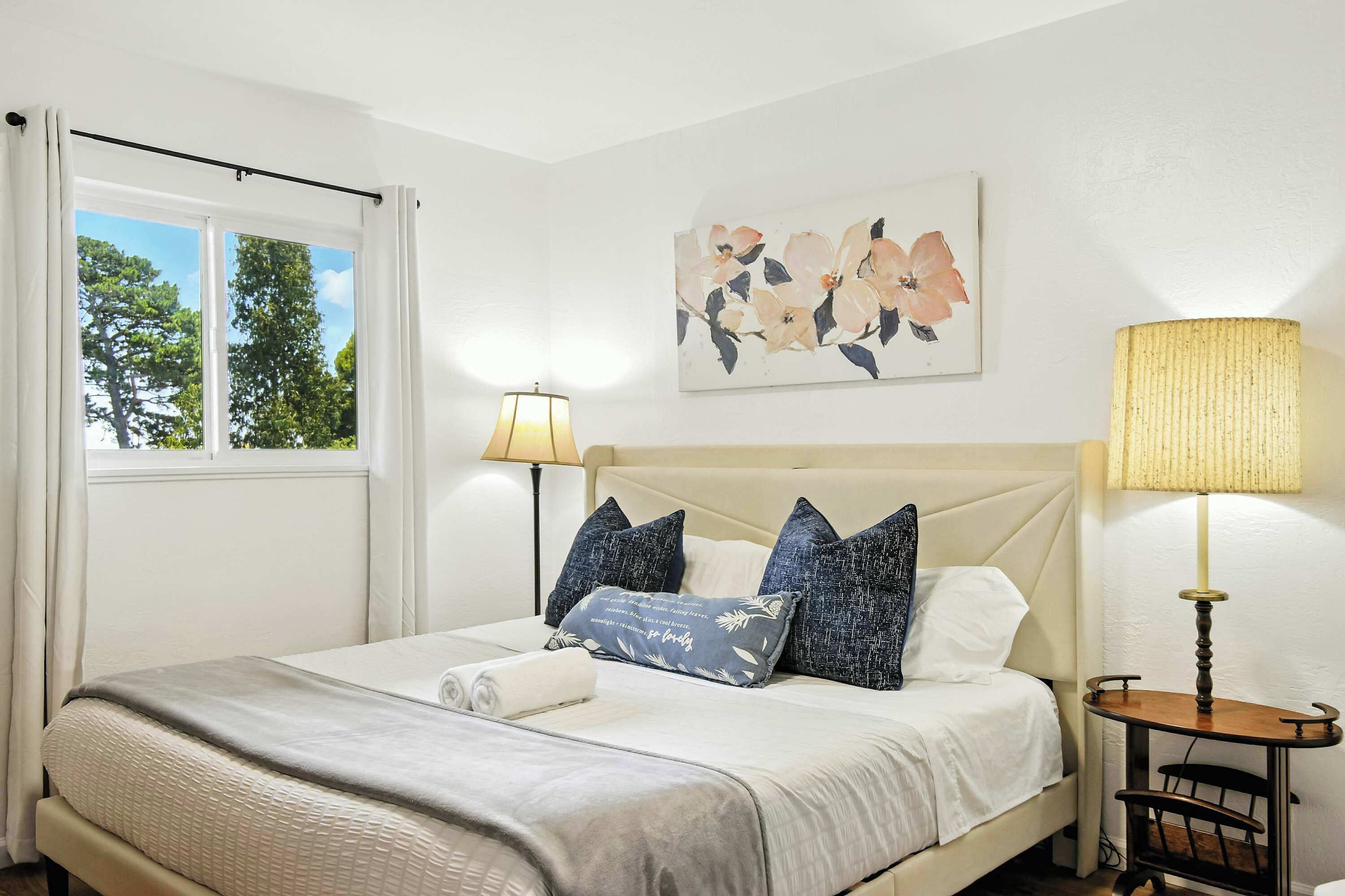 A neatly made bed with a white headboard, a small bedside table with a lamp, and a window with curtains.