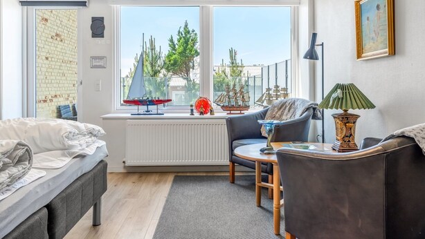 Ein modernes Wohnzimmer mit einem Bett, einem Sofa, einem Sessel und einem Tisch. Ein großes Fenster bietet Blick auf Bäume und ein Modell eines Segelbootes.